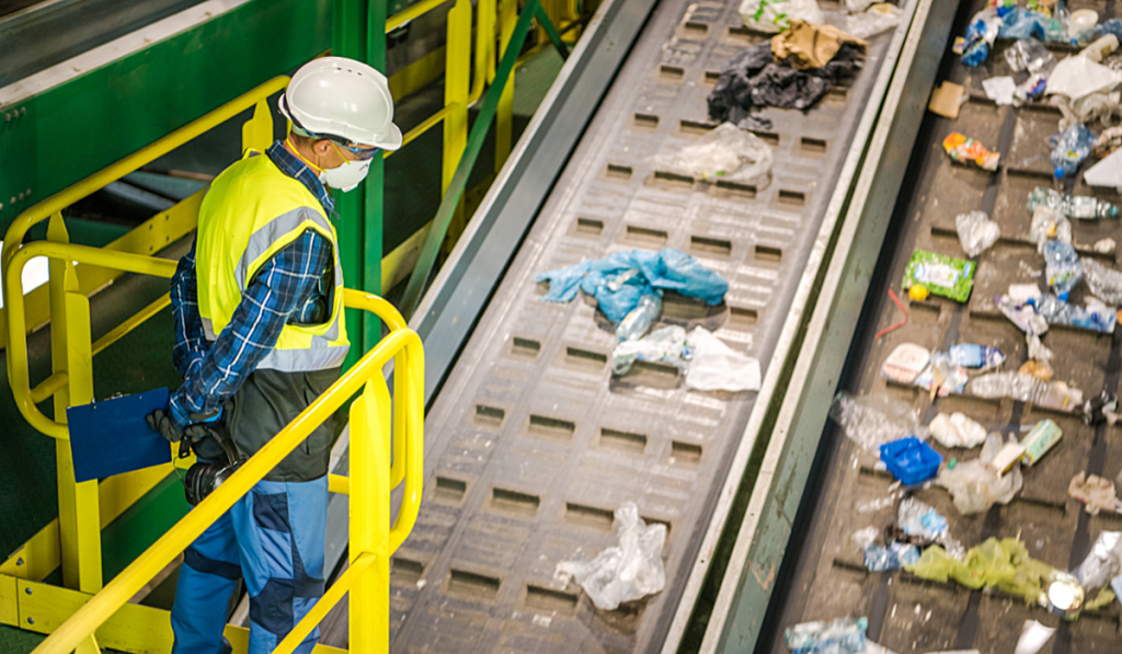 Worker monitoring waste sorting operations on a conveyor belt at a commercial waste management facility.