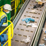 Worker monitoring waste sorting operations on a conveyor belt at a commercial waste management facility.