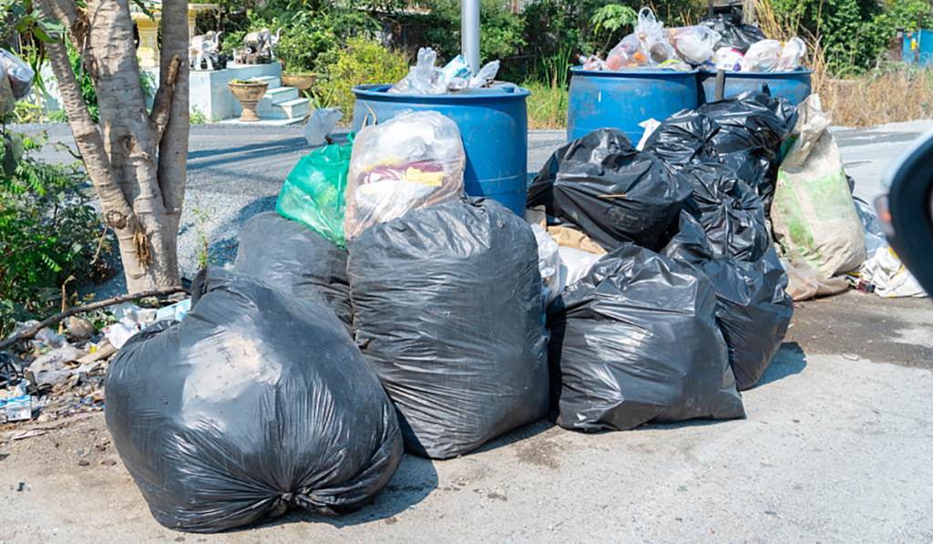 Black rubbish bags and blue waste bins filled with household rubbish outdoors.