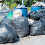 Black rubbish bags and blue waste bins filled with household rubbish outdoors.